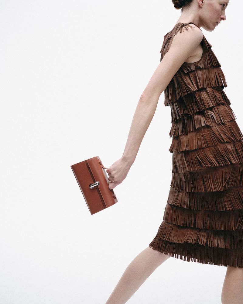 Fashion shot of a woman in a brown fringed dress holding a brown handbag against a white background in a studio setting. 