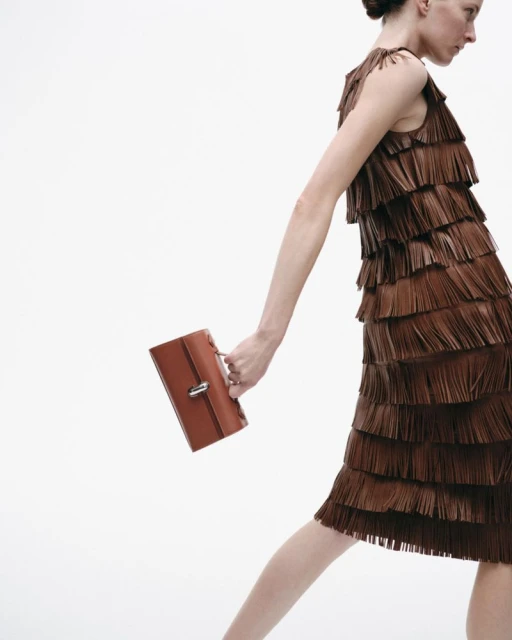 Fashion shot of a woman in a brown fringed dress holding a brown handbag against a white background in a studio setting. 