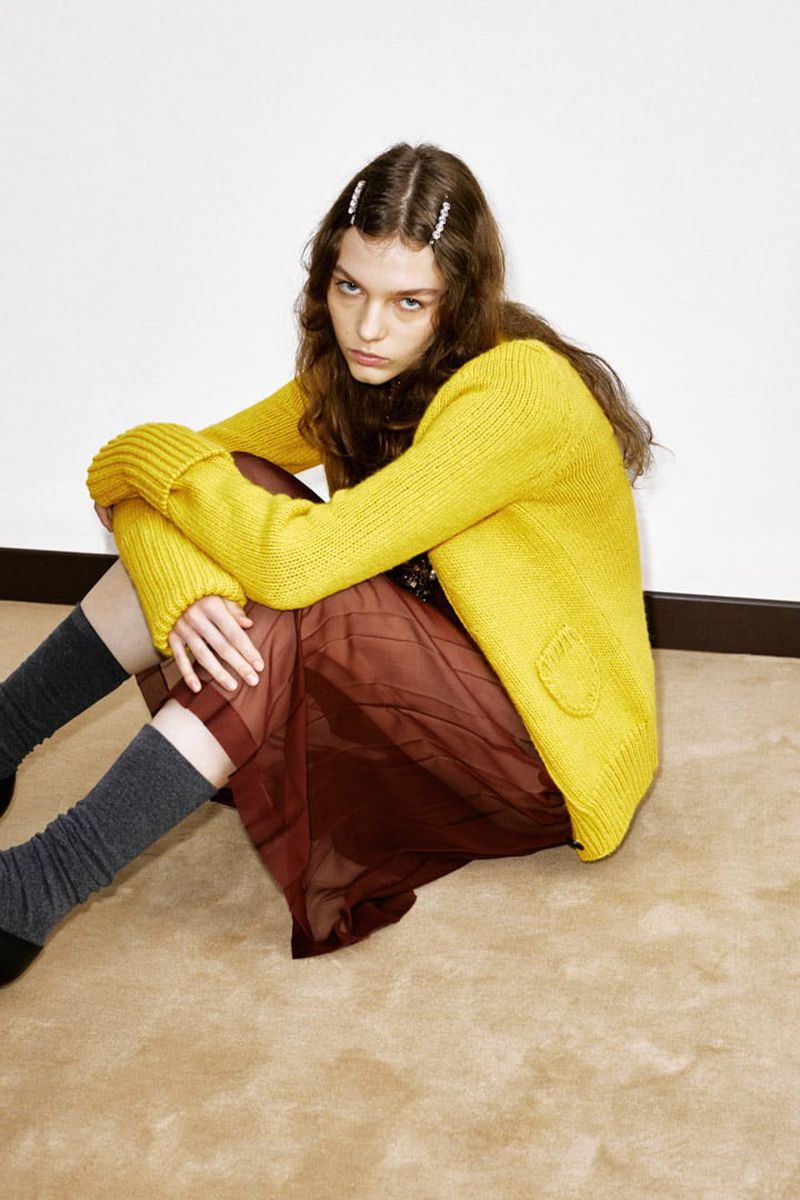 Young woman in a yellow cardigan and brown skirt sitting cross-legged on a beige floor with a white wall in the background.