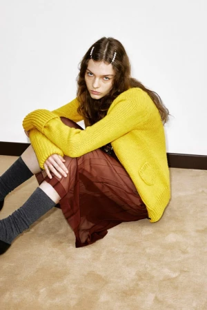 Young woman in a yellow cardigan and brown skirt sitting cross-legged on a beige floor with a white wall in the background.