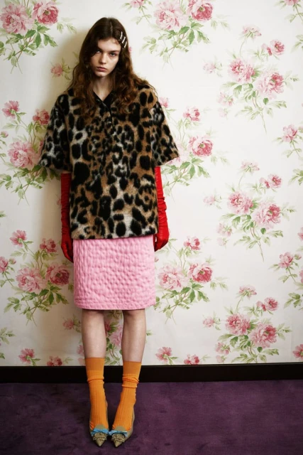 A slender young woman poses in a leopard print shirt and pink skirt against a floral backdrop, showcasing a colorful and stylish fashion ensemble.