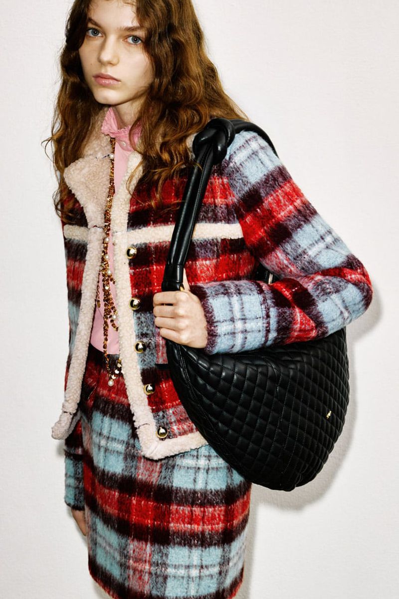 Fashion shot of a young woman wearing a red, white, and blue plaid skirt suit, holding a black, quilted leather hobo bag. 