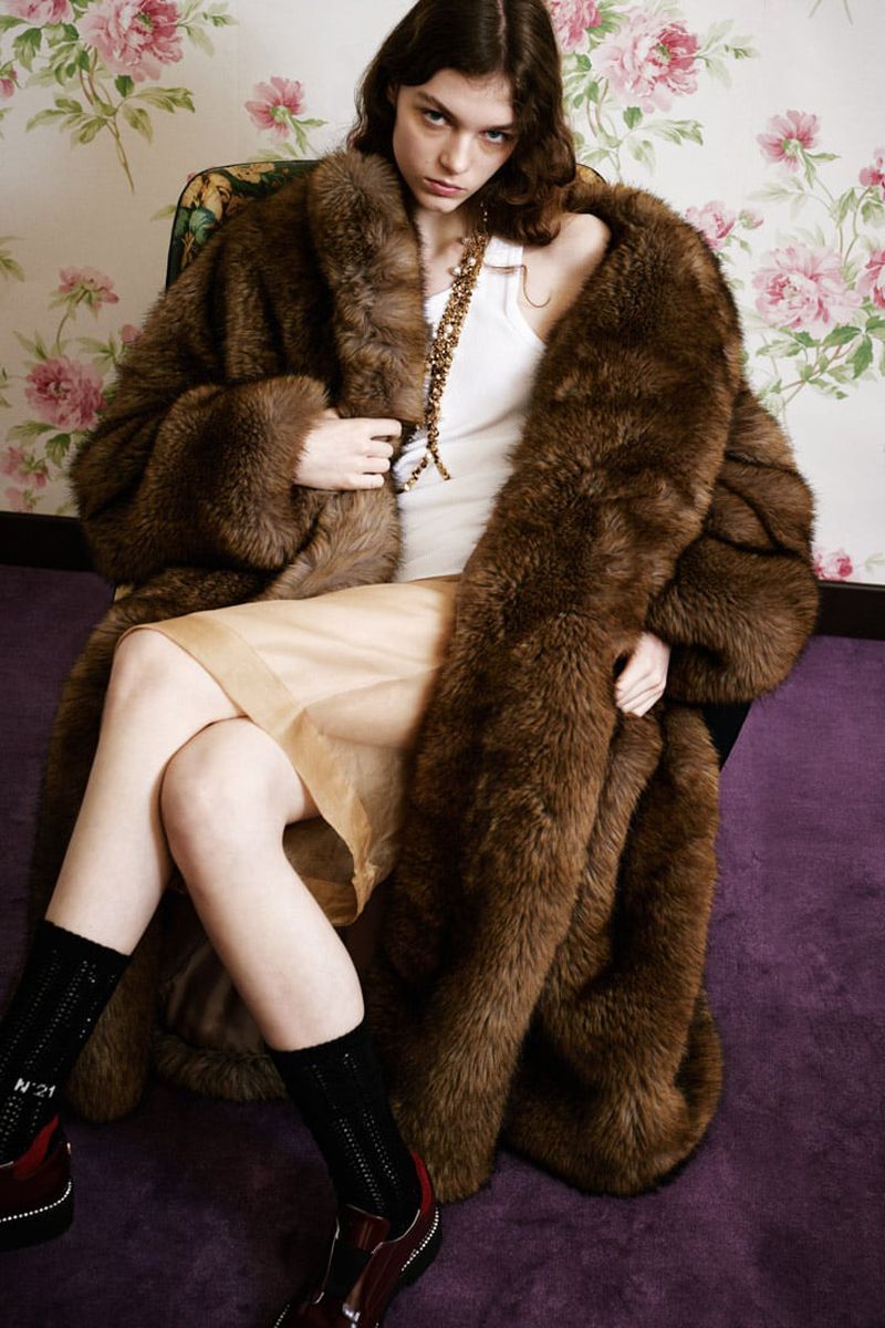Model wearing a large brown faux fur coat and cream skirt seated in an armchair against floral wallpaper.