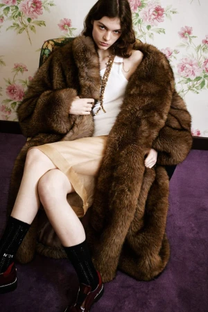 Model wearing a large brown faux fur coat and cream skirt seated in an armchair against floral wallpaper.
