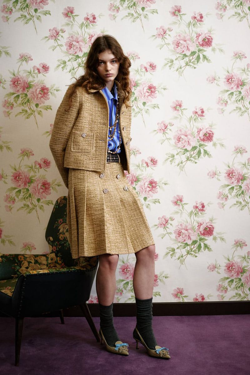 A young woman in a tweed skirt suit poses in front of a rose-patterned wall, with a floral chair visible at the left.