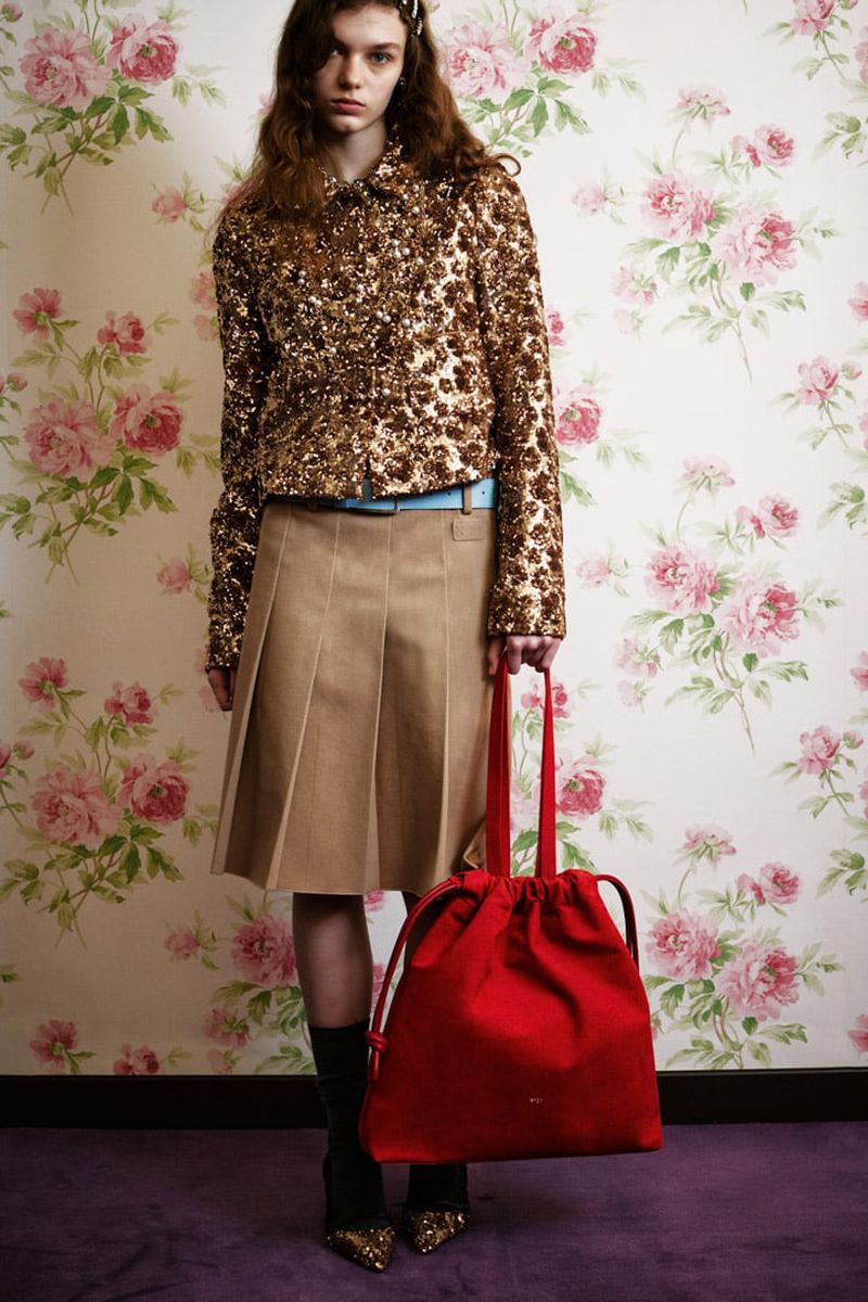 A woman poses in a sequined jacket and pleated skirt, holding a red bag against a floral wallpaper backdrop in a studio setting.