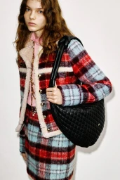 Model wearing a plaid jacket and matching skirt holding a black quilted handbag against a plain background