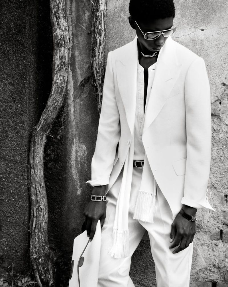 Black male model wearing an all-white suit and sunglasses, posing against a wall with exposed tree roots in a black and white photograph.