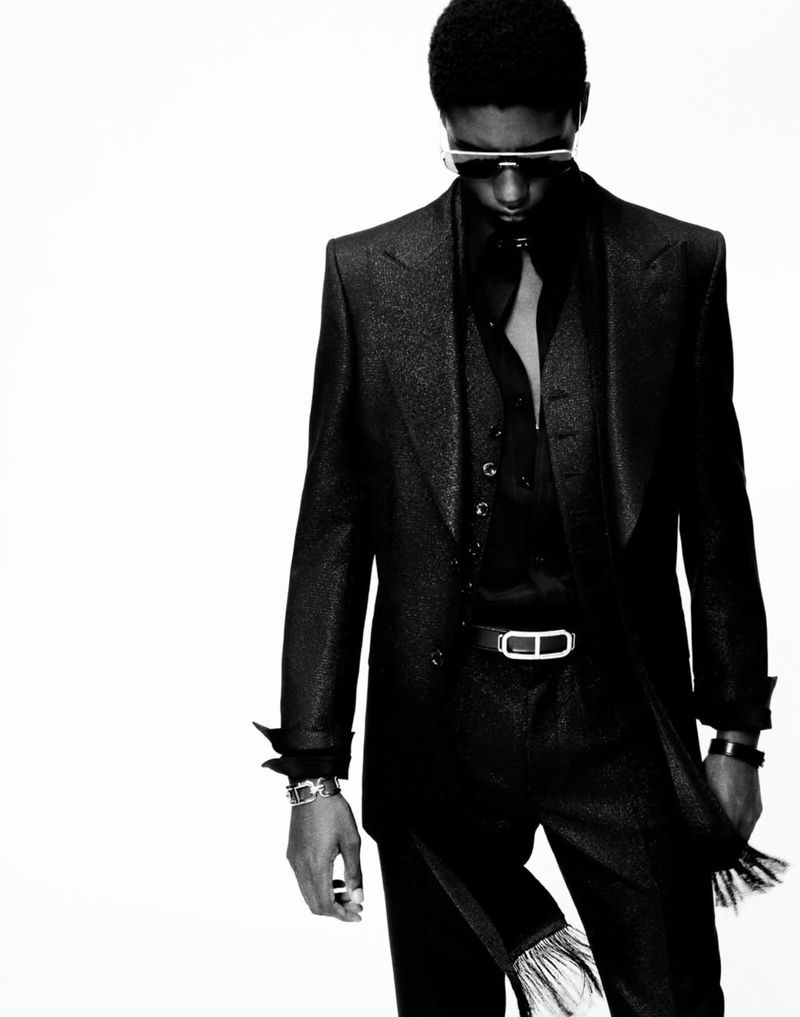 Black and white fashion shot of a slender adult male with an afro, wearing a dark sparkling suit and sunglasses against a white backdrop.