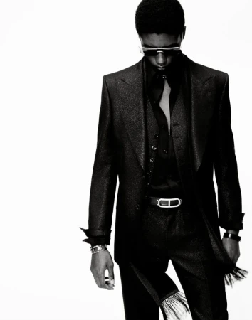 Black and white fashion shot of a slender adult male with an afro, wearing a dark sparkling suit and sunglasses against a white backdrop.