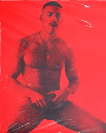 Male model with a mustache poses in a red room under plastic wrap. Man has a necklace, shaved head, and sits with his legs spread.