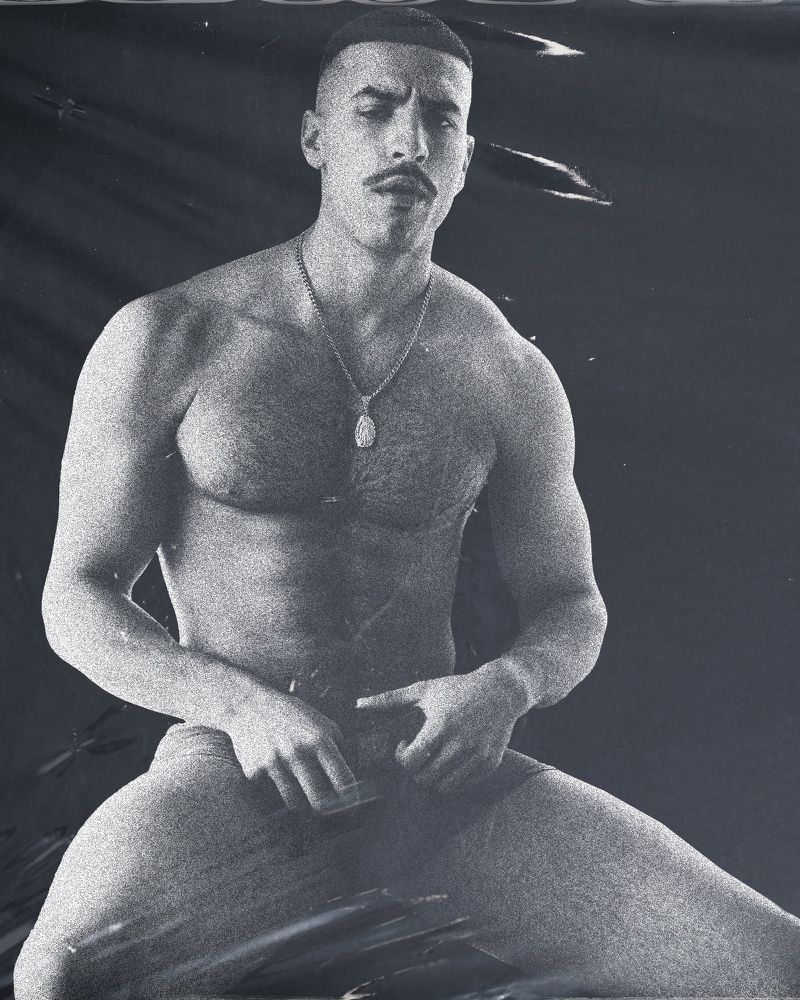 Portrait of a muscular, light-skinned adult man with a mustache, wearing a necklace, looking at the camera with a neutral expression.