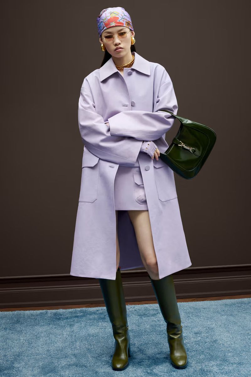 Fashion shot of a stylish young woman in lilac coat, skirt, olive boots, and patterned headscarf, holding a green leather purse.