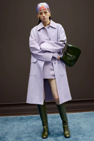 Fashion shot of a stylish young woman in lilac coat, skirt, olive boots, and patterned headscarf, holding a green leather purse.