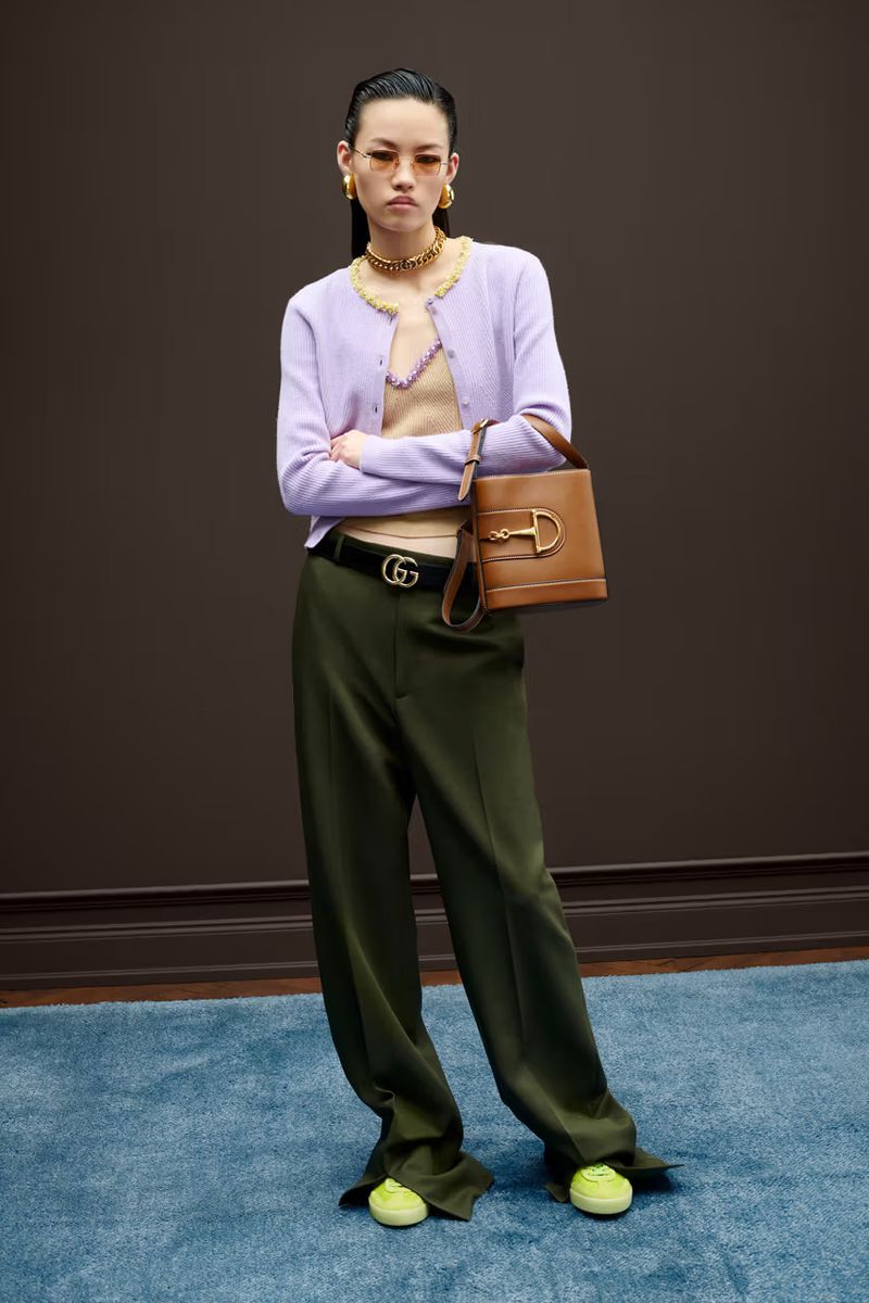 Full-shot of a woman in a lilac cardigan, beige top, olive pants, and sneakers, holding a tan bucket bag, standing in front of a brown wall.