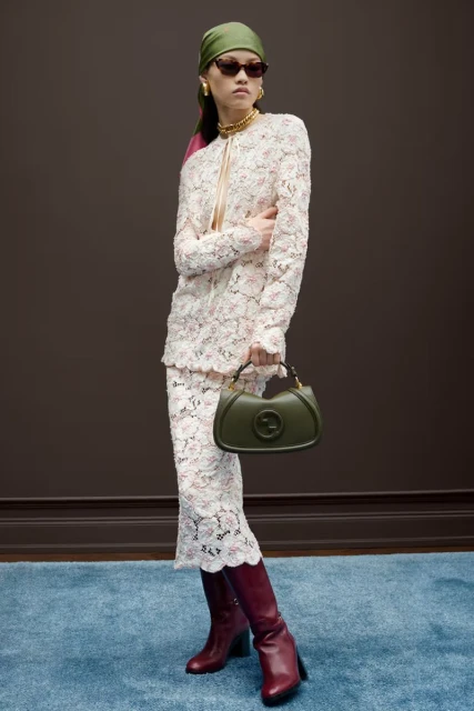 A woman poses in a lace skirt-suit, burgundy boots, headscarf, and sunglasses, holding a purse in a studio setting. Fashion editorial.