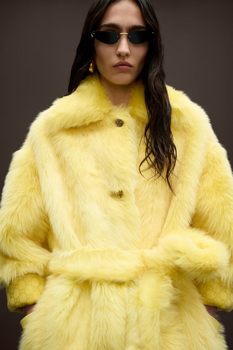 A brunette woman with sunglasses poses in a bright yellow faux-fur coat with a tie belt against a dark backdrop in a fashion shot.