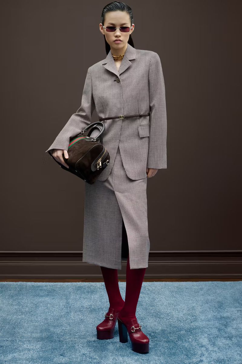 A slender Asian woman models a tailored skirt suit, paired with a dark handbag, red tights and platform shoes, against a brown backdrop.
