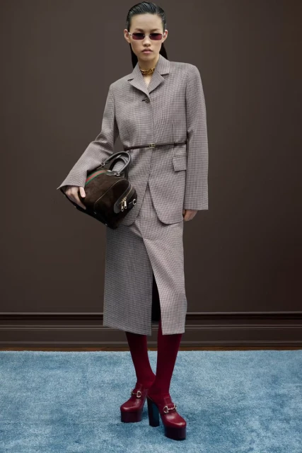 A slender Asian woman models a tailored skirt suit, paired with a dark handbag, red tights and platform shoes, against a brown backdrop.