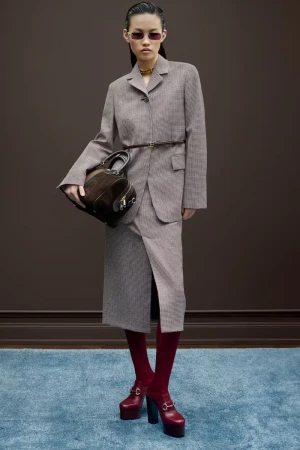 A slender Asian woman models a tailored skirt suit, paired with a dark handbag, red tights and platform shoes, against a brown backdrop.