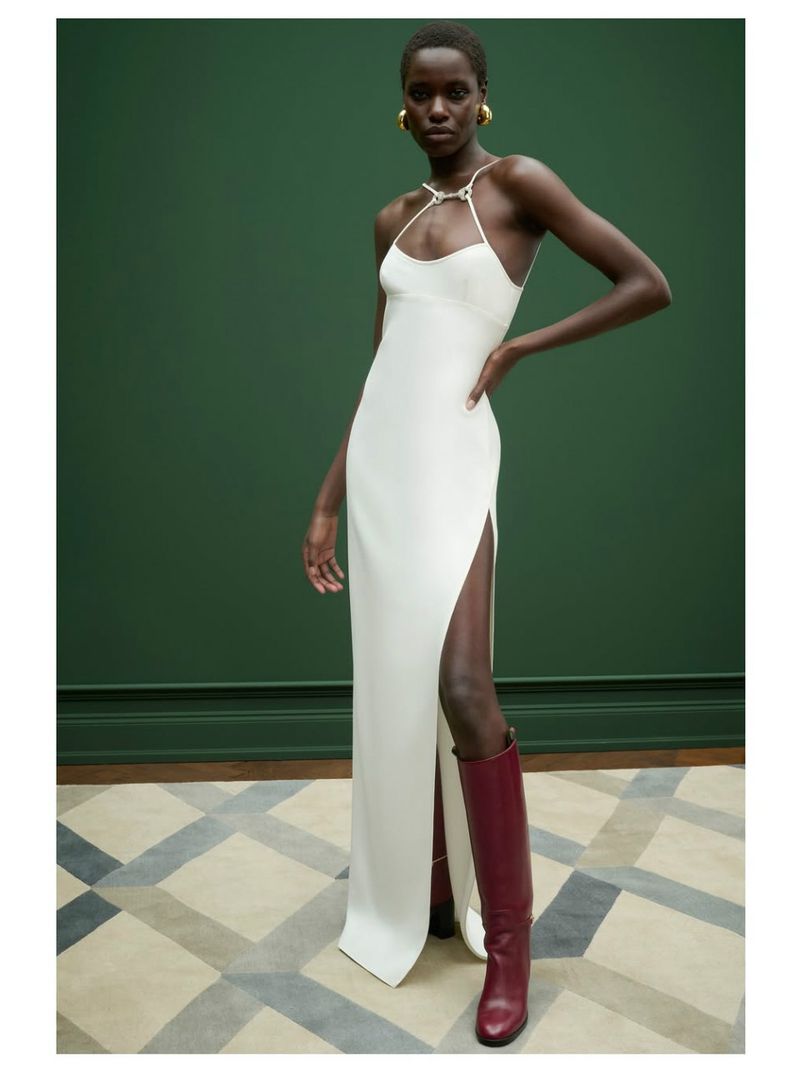 A dark-skinned female model poses in a long white dress with a slit and maroon knee-high boots against a green wall in a studio.