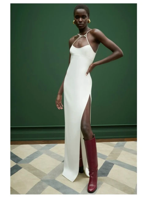 A dark-skinned female model poses in a long white dress with a slit and maroon knee-high boots against a green wall in a studio.