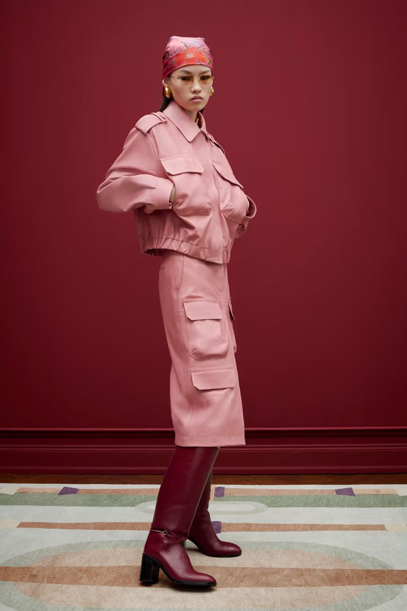 Studio shot of a stylish adult female in a matching pink jacket and skirt, accessorized with a headscarf, boots, and hoop earrings.