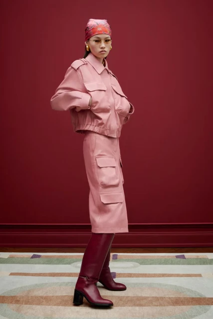 Studio shot of a stylish adult female in a matching pink jacket and skirt, accessorized with a headscarf, boots, and hoop earrings.