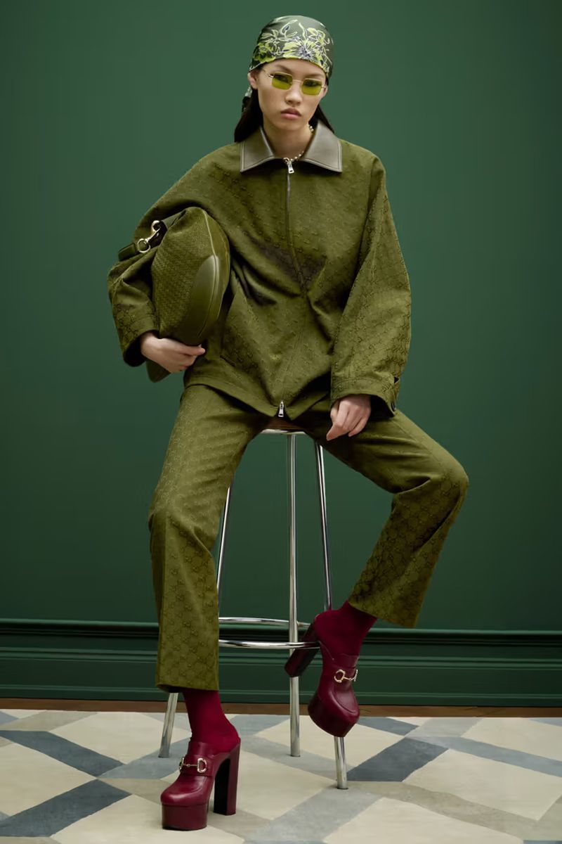 A woman in an olive-green suit sits on a stool, holding a matching bag, in a fashion studio with a geometric carpet and green backdrop.