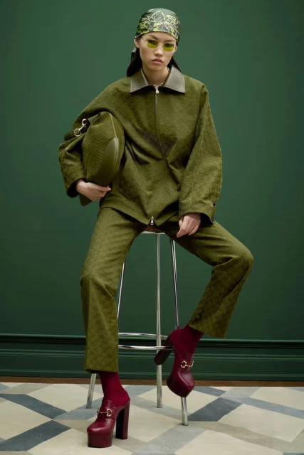 A woman in an olive-green suit sits on a stool, holding a matching bag, in a fashion studio with a geometric carpet and green backdrop.