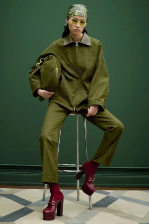 A woman in an olive-green suit sits on a stool, holding a matching bag, in a fashion studio with a geometric carpet and green backdrop.
