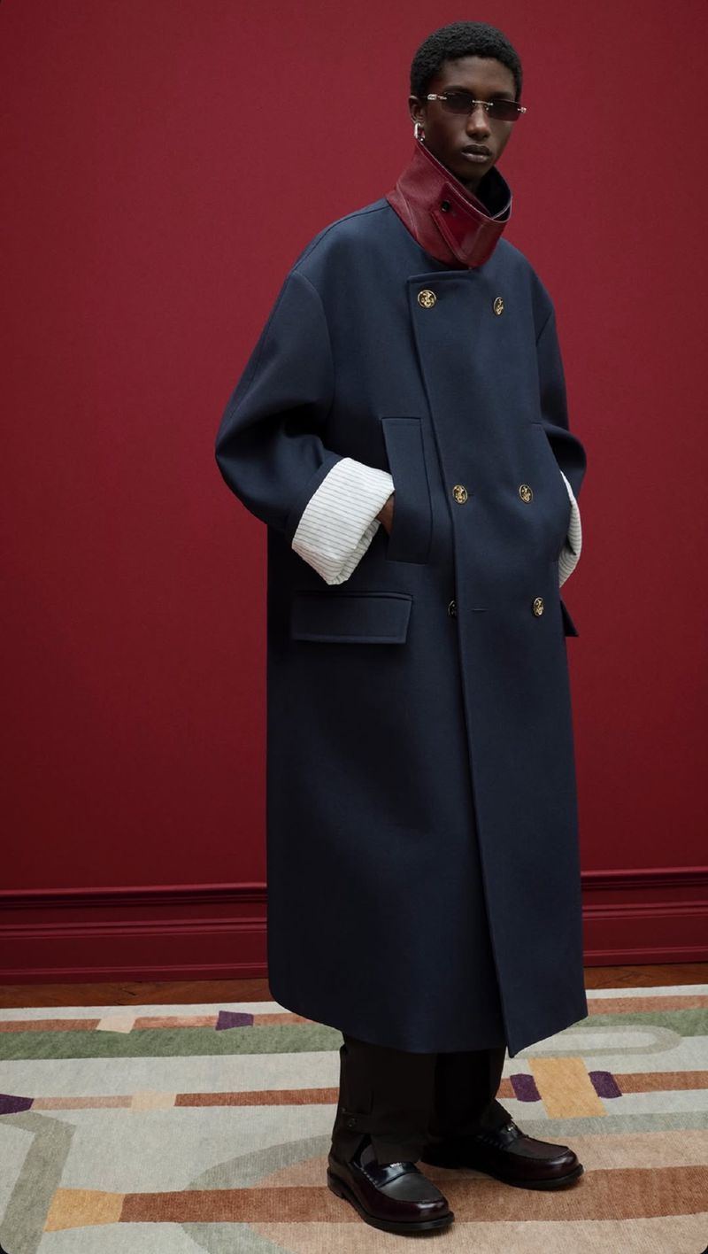 An adult male fashion model poses in a long blue double-breasted coat with gold buttons, dark pants and shoes against a red backdrop.
