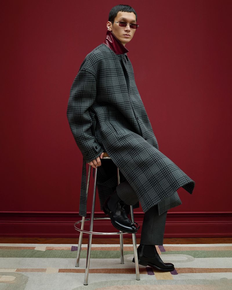 Stylish adult male sitting on a stool wearing a plaid coat, sunglasses, and black boots in front of a red wall indoors.