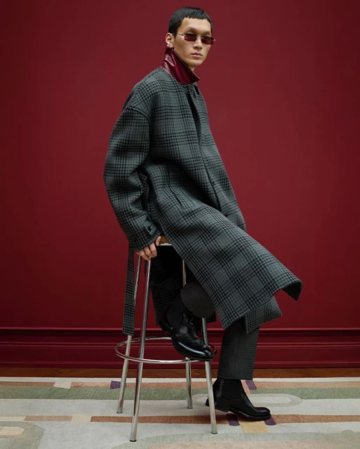 Stylish adult male sitting on a stool wearing a plaid coat, sunglasses, and black boots in front of a red wall indoors.