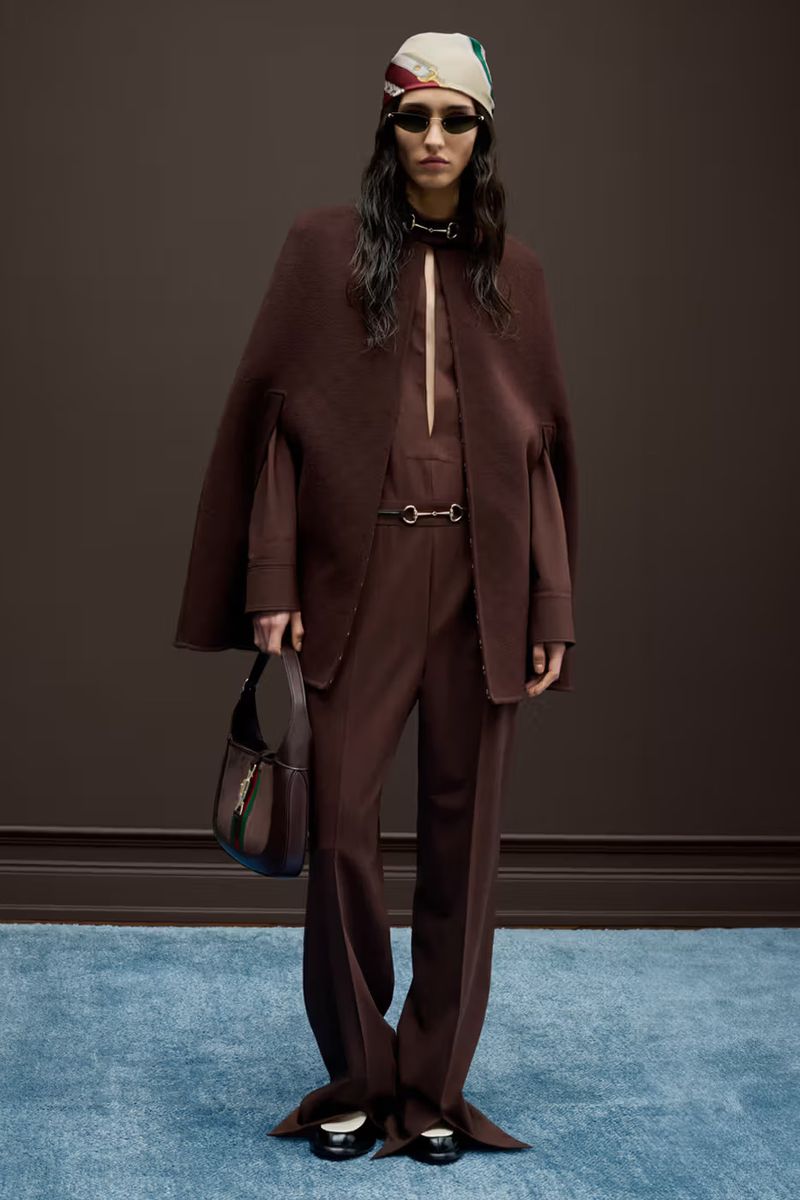Fashion model poses in brown cape and pants with matching purse, scarf and sunglasses in a studio shot for a fashion editorial.
