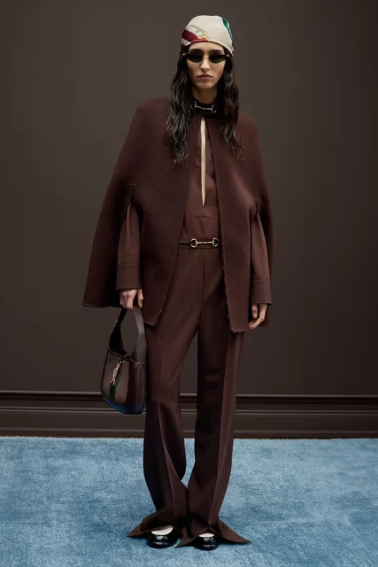 Fashion model poses in brown cape and pants with matching purse, scarf and sunglasses in a studio shot for a fashion editorial.
