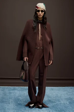 Fashion model poses in brown cape and pants with matching purse, scarf and sunglasses in a studio shot for a fashion editorial.