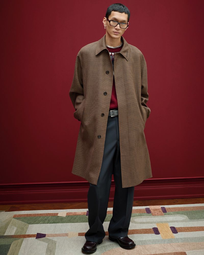 An Asian male model is posing in a brown plaid overcoat, sweater, and black dress pants in a fashion studio against a burgundy wall.