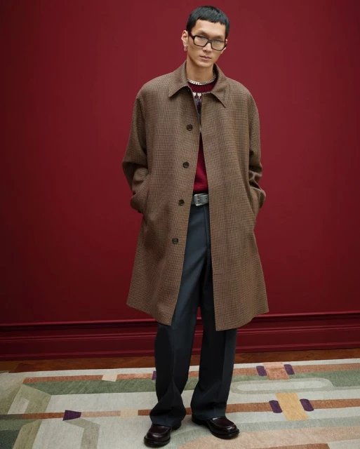 An Asian male model is posing in a brown plaid overcoat, sweater, and black dress pants in a fashion studio against a burgundy wall.