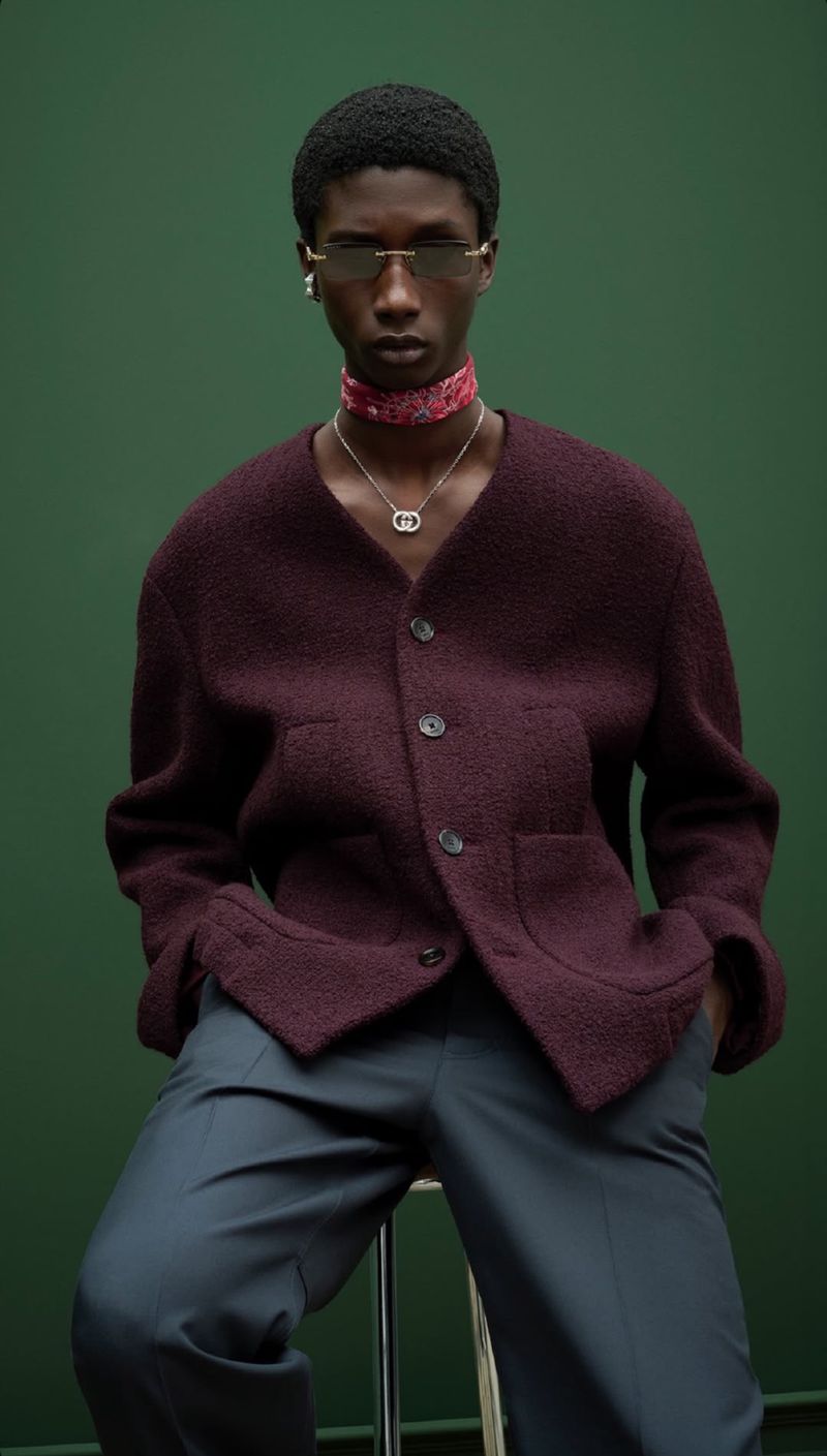 Studio portrait of a dark-skinned male model wearing a maroon jacket, gray pants, jewelry, and sunglasses against a green backdrop.