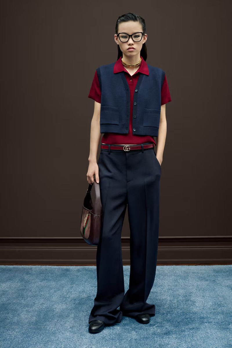 A stylish young woman models a navy vest and wide-leg trousers indoors on a mottled blue carpet, complemented by dark hair, glasses, and a purse.