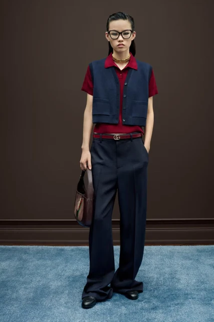 A stylish young woman models a navy vest and wide-leg trousers indoors on a mottled blue carpet, complemented by dark hair, glasses, and a purse.