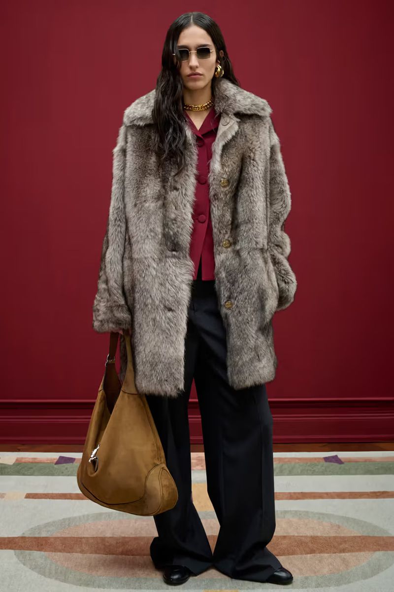Adult woman wearing a grey fur coat, maroon button-down shirt, black trousers, and sunglasses standing indoors with a brown handbag.