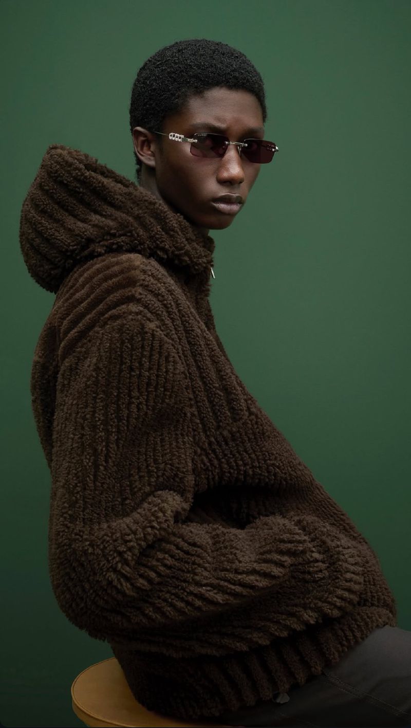 A young adult Black male with sunglasses wearing a brown, textured, hooded jacket, sits against a green backdrop in a studio setting.