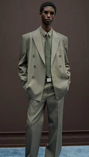 Model in olive green double-breasted suit and tie wearing sunglasses against brown wall