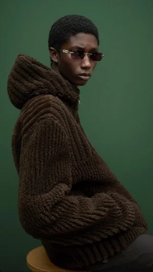 Model wearing a dark brown textured hooded jacket and sunglasses against a green background for Gucci Pre-Fall 2025 Lookbook