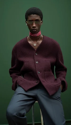 Black male model wearing sunglasses, red scarf, and a burgundy cardigan against a green seamless studio background