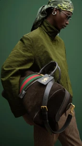 Model in olive green jacket holding brown leather duffel bag against dark green studio background