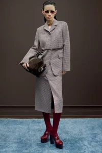 Model wearing a patterned suit jacket and skirt holding a handbag against a dark brown wall and blue carpet background.