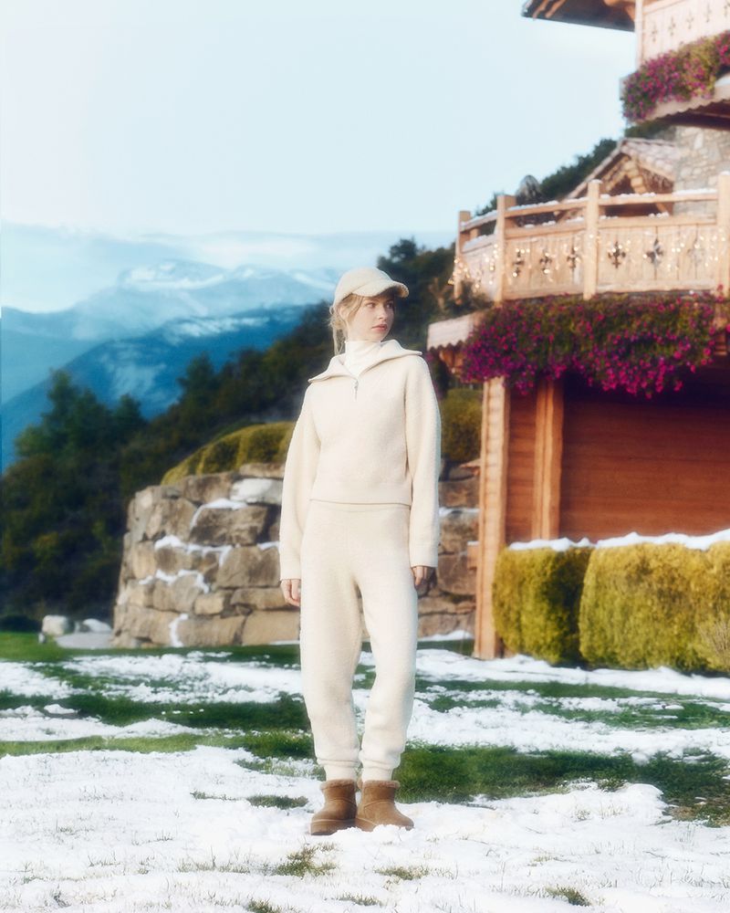 Blonde woman poses outdoors near house wearing beige sweater, sweatpants, cap, and boots in a snowy, mountainous landscape.